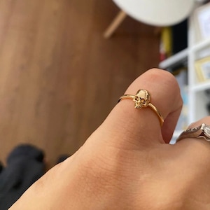 May include: A gold skull ring with a simple band. The ring is worn on a person's finger.