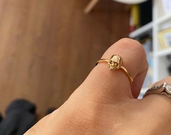May include: A gold skull ring with a simple band. The ring is worn on a person's finger.