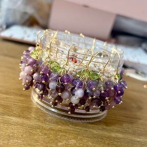 May include: A set of amethyst and rose quartz grape earrings with gold wire and green leaf accents. The earrings are displayed in a clear container.