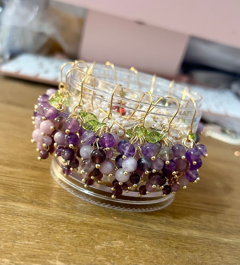 May include: A set of amethyst and rose quartz grape earrings with gold wire and green leaf accents. The earrings are displayed in a clear container.