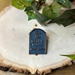 May include: Black enamel pin shaped like a tombstone with the text "Here Lies Ronnie Radke 1988 - 2023".