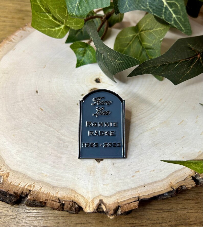 May include: Black enamel pin shaped like a tombstone with the text "Here Lies Ronnie Radke 1988 - 2023".
