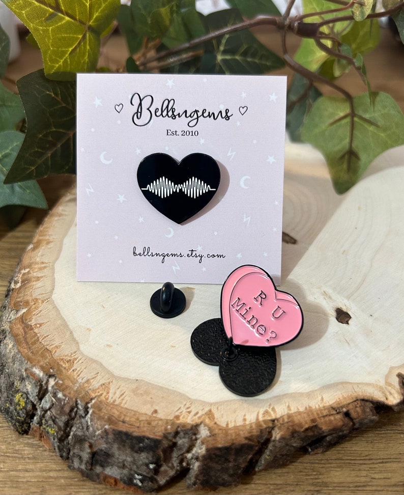 Puede incluir: Dos pines de esmalte en forma de coraz&oacute;n. El primer pin es negro y presenta una forma de onda estilizada dentro del coraz&oacute;n. El segundo pin es rosa y tiene el texto "RU Mine?" dentro del coraz&oacute;n.