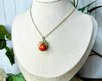 May include: A gold-colored chain necklace with a pendant shaped like a small orange pumpkin. The pumpkin pendant has green leaves and gold wire accents. The necklace is displayed on a white mannequin.