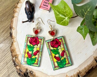 May include: A pair of gold earrings with red heart charms and stained glass rose designs. The roses are red with green leaves and stems. The earrings are on a wooden surface.