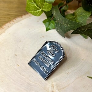 May include: A black metal pin shaped like a tombstone with the text "Here Lies Ronnie Radke 1983-2003".