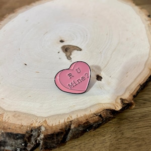 Puede incluir: Pin de esmalte en forma de coraz&oacute;n rosa con el texto "R U Mine?" sobre una superficie de madera.