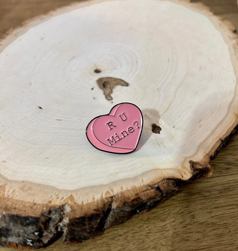 Puede incluir: Pin de esmalte en forma de coraz&oacute;n rosa con el texto "R U Mine?" sobre una superficie de madera.