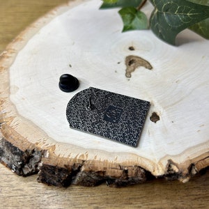 May include: Black enamel pin with a rounded rectangular shape. The pin has a textured surface and a small, round, black pin back.