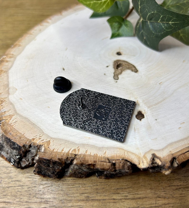 May include: Black enamel pin with a rounded rectangular shape. The pin has a textured surface and a small, round, black pin back.