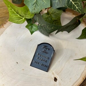 May include: Black enamel pin shaped like a tombstone with the text "Here Lies Ronnie Radke 1983-2033".