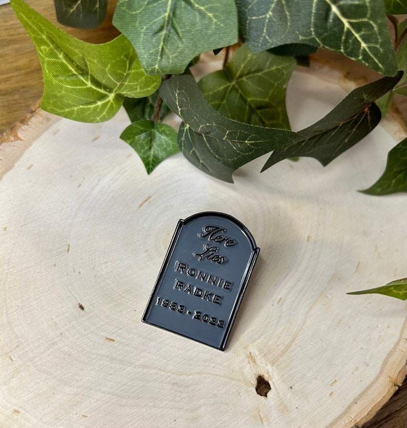 May include: Black enamel pin shaped like a tombstone with the text "Here Lies Ronnie Radke 1983-2033".