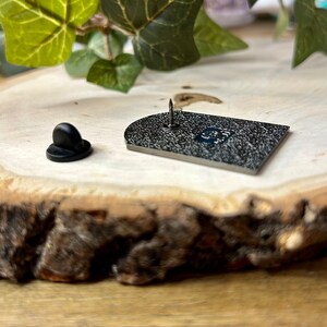 May include: The back of a black enamel pin with a silver pin back. The pin is shaped like a rectangle with rounded corners. The pin is on a wooden surface.