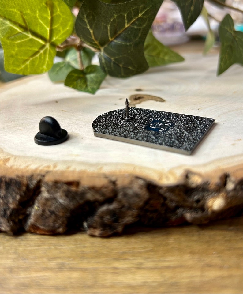 May include: The back of a black enamel pin with a silver pin back. The pin is shaped like a rectangle with rounded corners. The pin is on a wooden surface.