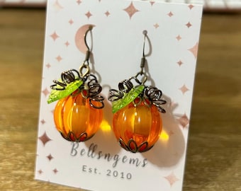 May include: A pair of orange pumpkin earrings with green leaf accents and bronze-colored metalwork. The earrings are displayed on a white card with the text "Bellsngems Est. 2010" and a pattern of pink stars.