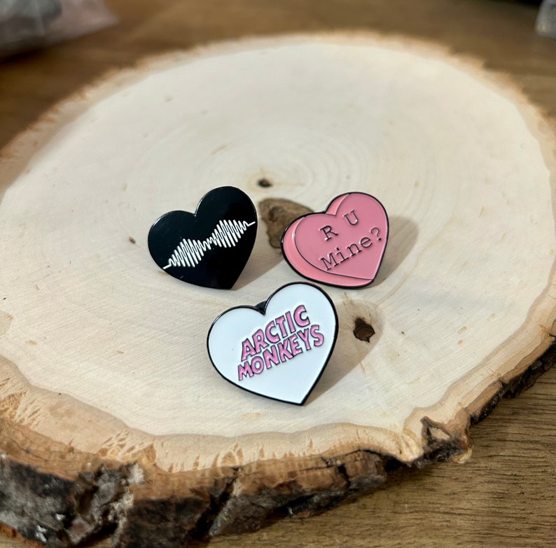 Puede incluir: Tres pines en forma de coraz&oacute;n con esmalte negro, blanco y rosa. El pin negro tiene un patr&oacute;n de onda blanco en el interior. El pin blanco tiene las palabras "Arctic Monkeys" en letras rosas. El pin rosa tiene las palabras "R U Mine?" en letras blancas.