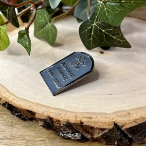 May include: A silver-coloured enamel pin shaped like a tombstone with the text "Here Lies Ronnie Radke 1988-2023".