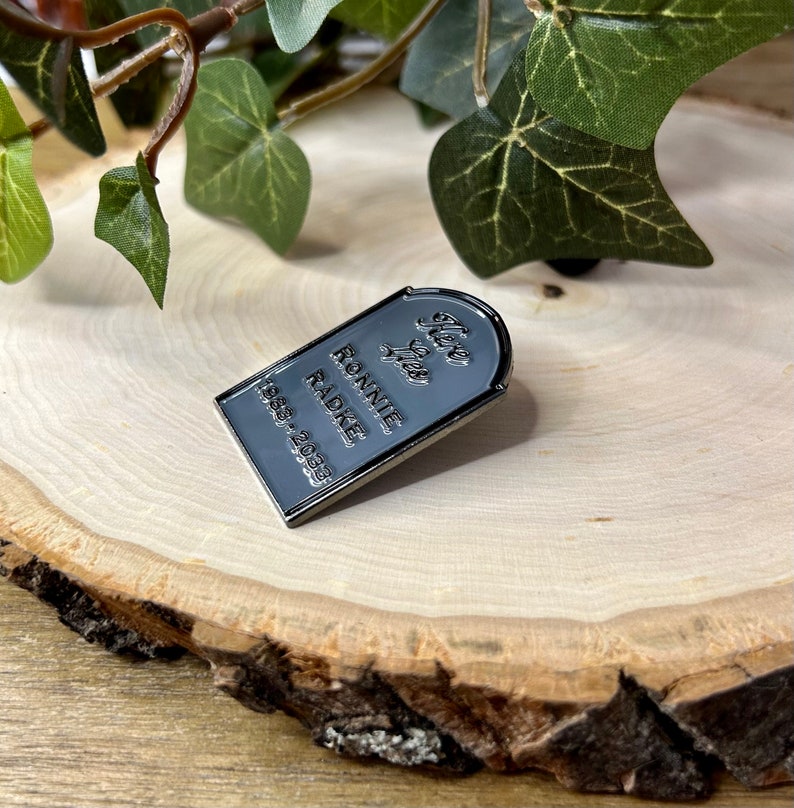 May include: A silver-coloured enamel pin shaped like a tombstone with the text "Here Lies Ronnie Radke 1988-2023".