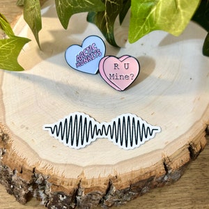 May include: A collection of music-themed accessories, including two heart-shaped pins and a sticker. The pins feature the text "Arctic Monkeys" and "R U Mine?" on a light blue and pink background. The sticker displays a black and white sound wave design.