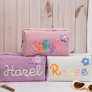 Può includere: Tre trousse per cosmetici in velluto a coste rosa, lavanda e bianco. Ogni borsa ha un nome ricamato con filo colorato, con abbellimenti floreali. La borsa rosa dice "Sibyl", la borsa lavanda dice "Harel" e la borsa bianca dice "Renee".