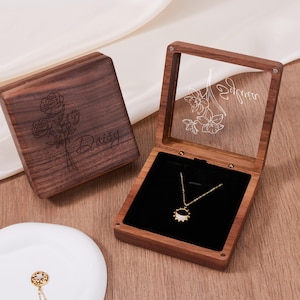 May include: A wooden jewelry box with a clear lid, showcasing a gold necklace with a sunburst pendant. The box is open, revealing a black velvet interior. The lid features a floral design and the name "Adrina". The closed box has the name "Daisy" and a floral design.