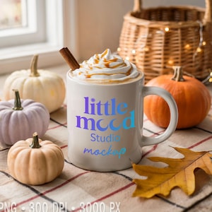 Op de afbeelding: Een witte mok met de tekst "little mood Studio mockup" bevat een drankje gegarneerd met slagroom, karamel en een kaneelstokje. Kleine pompoenen in verschillende kleuren omringen de mok, met een rieten mand op de achtergrond.