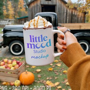 Puede incluir: Una taza blanca con el texto "little mood Studio mockup" en morado y azul, llena de crema batida y caramelo. La taza se sostiene frente a un camión negro, una caja de madera con manzanas y una calabaza.