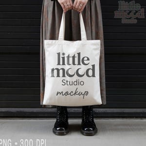Może przedstawiać: Biała torba typu tote z napisem "little mood Studio mockup" w ciemnoszarym kolorze. Torba jest trzymana przez osobę noszącą długą spódnicę i czarne buty. Tło to ciemnoszara ściana.