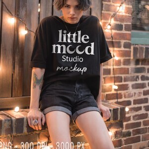 May include: Black t-shirt with the text "little mood Studio mockup" in white. The model is wearing black denim shorts. The background includes a brick wall and string lights.