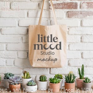 May include: A beige canvas tote bag with the text "little mood studio mockup" in dark gray. The bag hangs in front of a white brick wall. A variety of potted succulents are arranged on a wooden shelf below the bag.