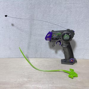 以下が含まれることがあります： おもちゃの乗り物用のグレーと黒のピストル型リモコン。紫のアクセント、長く細い金属製アンテナ、27MHZと書かれた緑色の円形ラベルが付いています。別個の柔軟な明るい緑色のプラスチック製ギザギザのプルコードアクセサリーが付属しています。