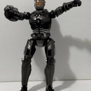 Puede incluir: Una figura de acción articulada de un personaje robótico con armadura gris metálico y detalles en negro. La figura cuenta con un casco con visera naranja translúcida, extremidades articuladas y una placa pectoral con un pequeño emblema rojo.