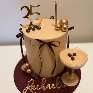 May include: A coffee-themed birthday cake with tan frosting, decorated with gold accents. The cake has a gold "35" topper, a candle, and chocolate-covered coffee beans. A small cocktail glass with coffee cream sits beside the cake. The name "Rachael" is written in gold.