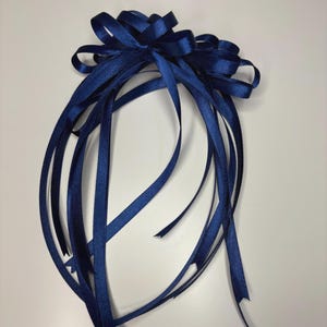 May include: A navy blue satin ribbon bow with long, flowing streamers. The bow is tied at the top and has multiple loops. The ribbons are a deep, rich blue colour and are arranged against a white background.