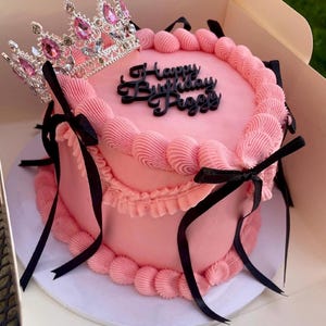 May include: A round pink cake decorated with pink piped frosting borders, black ribbon bows, and a silver-toned crown with pink gems on top. The cake features a black topper with the text Happy Birthday Peggy in a cursive script font.