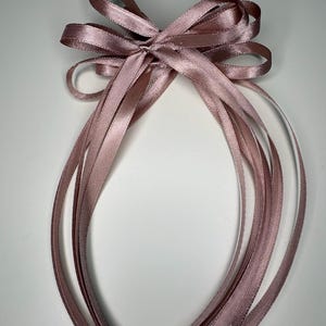 May include: A dusky pink satin ribbon bow with long, flowing tails. The bow is tied in the centre and has multiple loops. The ribbon has a slight sheen, and the ends of the tails are cut at an angle. The background is white.