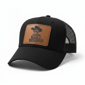 May include: Black trucker hat with a mesh back and a curved brim. The hat features a brown rectangular patch with a skull wearing a cowboy hat and the words "BADLY BEHAVED" in a bold font.