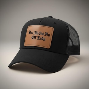 May include: Black trucker hat with a mesh back and a curved brim. A rectangular brown leather patch on the front displays the text "Let Me Ask My Ol' Lady" in a gothic font. The hat is a classic accessory.