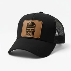 May include: Black trucker hat with a mesh back and a curved brim. A brown leather patch on the front features a military helmet, American flag, and the words "SUPPORT OUR TROOPS" in black.