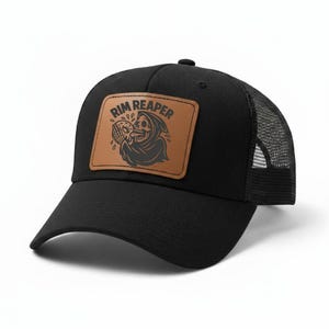 May include: Black baseball cap with a mesh back and a curved brim. The cap features a brown leather patch with the words "Rim Reaper" and a graphic of a grim reaper holding a skull. The cap is a trucker style.