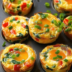 May include: A dark gray muffin tin containing several baked egg bites. Each round egg bite features a golden yellow cooked egg base mixed with bright red bell pepper pieces, dark green spinach, and topped with fresh chopped green onion slices.