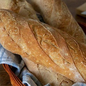 Peut inclure: Deux baguettes dorées à la croûte croustillante et incisée, disposées dans un panier en osier tressé doublé d'un tissu à carreaux bleus et blancs. Le pain présente une forme allongée rustique avec de la farine sur la surface texturée.