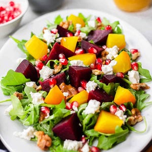 Peut inclure: Une salade sur une assiette blanche composée de morceaux de betteraves rouge foncé et jaune vif, de feuilles de roquette vertes, de fromage blanc émietté, de graines de grenade et de noix, offrant un mélange coloré de textures variées.