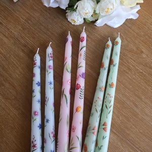 Peut inclure: Une collection de minces bougies coniques à motifs floraux, dans les tons rose, vert et blanc. Les bougies sont ornées de fleurs et de feuillages délicats peints à la main. Les bougies sont disposées sur une surface en bois, avec des fleurs blanches en arrière-plan.