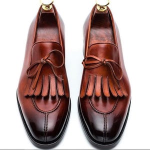 Op de afbeelding: Een paar bruine leren tassel loafers met een spitse neus en een franje detail. De loafers hebben een gepolijste afwerking en een subtiel ombre-effect.