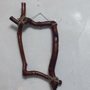 May include: A rustic, handcrafted wall hanging made from dark brown branches. The branches are joined with twine to form a unique, abstract shape. The piece is suspended by a thin wire, showcasing a natural, organic aesthetic. The wall decor is mounted on a white wall.