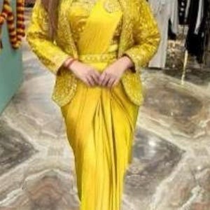 May include: A person wearing a vibrant yellow outfit, comprising a draped dress and a jacket with detailed embroidery. The outfit is complemented by a silver necklace. The setting appears to be a clothing boutique.