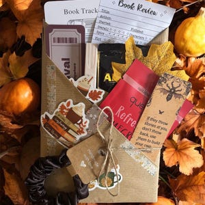 May include: A brown paper gift package containing a book review card, a bookmark with a beetle illustration and text, a black scrunchie, stickers, and a red tea packet. The package is tied with twine and features a tag labelled Handmade with love.