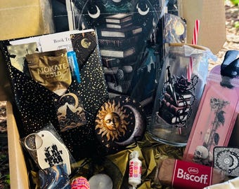 May include: A gift box containing a book-themed pouch, a glass tumbler with a straw, a Biscoff biscuit, a Mokate Gold coffee packet, a sun and moon coaster, a keychain with the text In My Fantasy Era, a bookmark with a skull, and assorted small treats.