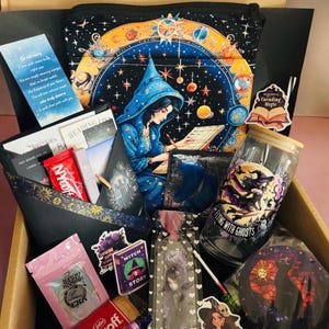 May include: A gift box containing a blue wizard-themed zippered pouch, a glass tumbler with a witch graphic and the text Flying with Ghosts, a bookmark, stickers, a card with text, a small book-shaped item, and various snacks in packaging.